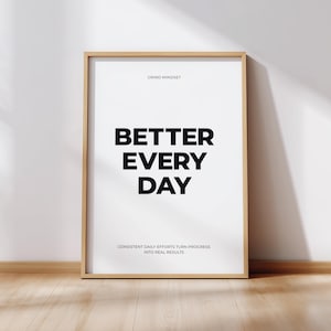 May include: A framed print with the words "BETTER EVERY DAY" in bold black letters on a white background. The phrase "GRIND MINDSET" is above the main text, and a motivational quote is below. The frame is a light wood color.