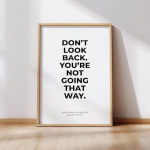 Pode incluir: Uma impressão emoldurada com o texto "DON'T LOOK BACK. YOU'RE NOT GOING THAT WAY." em letras pretas em negrito sobre um fundo branco. A parte inferior da impressão diz "EVERYTHING YOU NEED IS AHEAD OF YOU." A moldura é de cor castanho claro.
