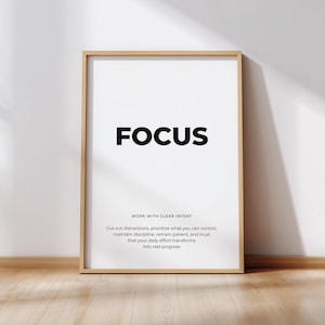 Peut inclure: Une affiche encadrée avec le mot « FOCUS » en lettres noires en gras. En dessous, un texte plus petit indique « WORK WITH CLEAR INTENT » et un message de motivation. Le cadre est marron clair et le fond est blanc.