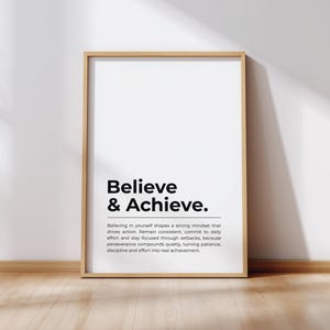 Pode incluir: Uma impressão emoldurada com as palavras "Believe & Achieve" em negrito preto. Abaixo do título há um parágrafo de texto. A moldura é marrom claro, e o fundo é uma parede branca e um chão de madeira.