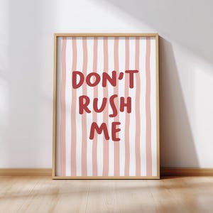 May include: A framed art print with a light wood frame. The artwork features a white background with vertical pink stripes. Bold red text in the center reads "DON'T RUSH ME". The print is displayed on a wooden floor against a white wall.