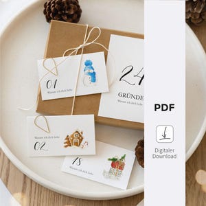 Op de afbeelding: Een bruine geschenkdoos met witte kaarten met aquarel illustraties en nummers. De kaarten zijn bevestigd met gouden paperclips en touw. De kaarten hebben illustraties van een sneeuwpop, peperkoekhuisje en cadeaus. De tekst op de kaarten luidt "Warum ich dich liebe".