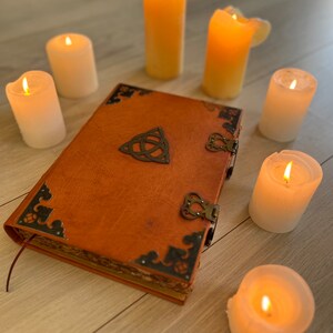 May include: A brown leather-bound journal with metal accents and a Celtic knot design on the cover. The journal is surrounded by eight lit candles on a wooden surface.
