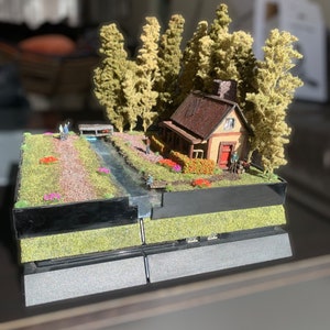 Puede incluir: Un diorama en miniatura de una pequeña cabaña con una puerta roja, rodeada de árboles y un arroyo, construida sobre una consola PlayStation 4 negra.