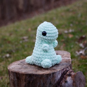 May include: A light green crocheted dinosaur toy with black eyes. The toy is sitting on a tree stump.