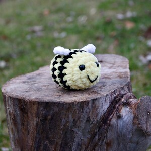 May include: A crocheted bee plush toy with black and yellow stripes, white wings, and a smiling face. The bee is sitting on a tree stump.