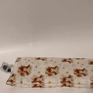 May include: A rectangular eye pillow with a white background and a repeating pattern of brown Highland cow faces and yellow flowers. A small tag is attached to the pillow. The pillow is designed for relaxation and comfort.