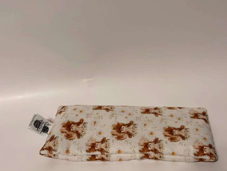 May include: A rectangular eye pillow with a white background and a repeating pattern of brown Highland cow faces and yellow flowers. A small tag is attached to the pillow. The pillow is designed for relaxation and comfort.