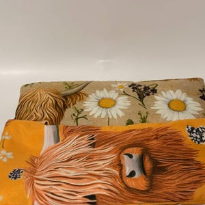 May include: Two rectangular fabric items with Highland cow and daisy patterns. One has an orange background with a cow's face, the other has a tan background with daisies. Both have a tag with text.