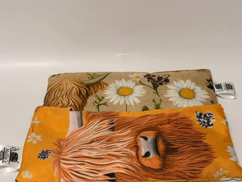 May include: Two rectangular fabric items with Highland cow and daisy patterns. One has an orange background with a cow's face, the other has a tan background with daisies. Both have a tag with text.