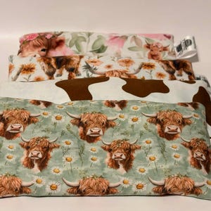 May include: A stack of four rectangular fabric items with various cow-themed patterns. The patterns include Highland cows, floral designs, and a cowhide print. The bottom item features Highland cows and daisies on a green background.