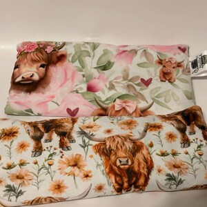May include: Two rectangular pillows with Highland cow designs. One pillow features a cow with a floral crown and pink accents. The other pillow has a cow with orange flowers. Both pillows have a white background.