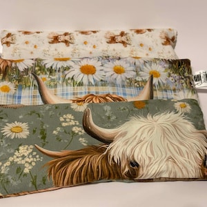 May include: Three decorative pillows with different floral and animal patterns. The top pillow has a cream background with brown bears. The middle pillow has large daisies on a blue plaid background. The bottom pillow features a Highland cow.