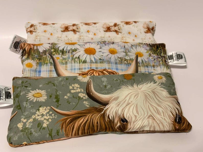 May include: Three decorative pillows with different floral and animal patterns. The top pillow has a cream background with brown bears. The middle pillow has large daisies on a blue plaid background. The bottom pillow features a Highland cow.