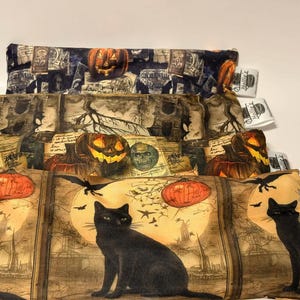 May include: Stacked Halloween-themed fabric pouches with various designs. The top pouch features a pumpkin and tombstone pattern. The middle pouch has a pumpkin, skull, and tree design. The bottom pouch showcases a black cat, bats, and a full moon.