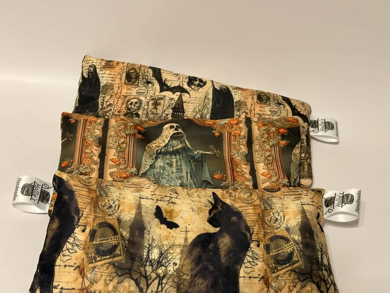 May include: Three Halloween-themed fabric pouches stacked on top of each other. The top pouch features a design with skulls and bats. The middle pouch has a ghost design. The bottom pouch has a black cat design.