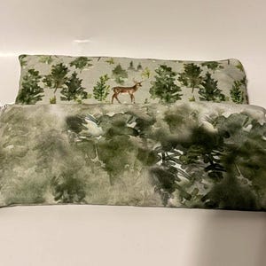 May include: Two rectangular throw pillows. The top pillow has a light gray background with a forest scene featuring green trees and a deer. The bottom pillow has a watercolor-style forest design in shades of green and gray.