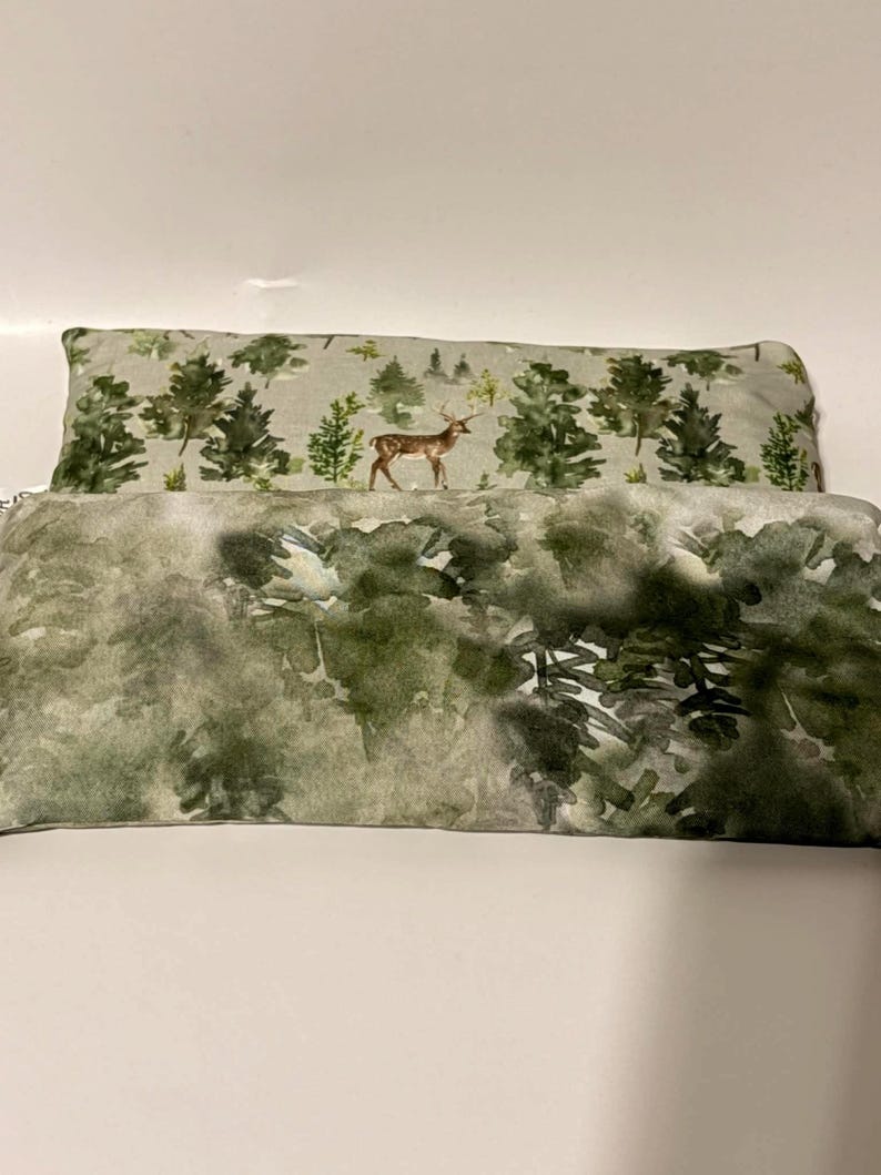 May include: Two rectangular throw pillows. The top pillow has a light gray background with a forest scene featuring green trees and a deer. The bottom pillow has a watercolor-style forest design in shades of green and gray.