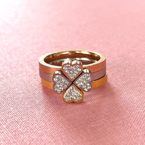 Puede incluir: Tres anillos apilados en oro, plata y oro rosa. El anillo superior presenta un diseño de trébol de cuatro hojas con piedras de circonita cúbica transparentes.