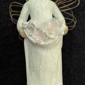 Puede incluir: Una figurita de porcelana blanca de un ángel sosteniendo un ramo de flores rosas. El ángel tiene el pelo rubio y lleva un vestido blanco.