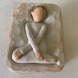 Könnte beinhalten: Eine kleine, weiße Holzfigur eines sitzenden Menschen mit gekreuzten Beinen. Die Figur befindet sich in einem rechteckigen Rahmen mit einer strukturierten Oberfläche.