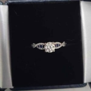 May include: A diamond engagement ring with a large, round center stone and smaller accent stones. The ring features a twisted band design with blue sapphire accents, set in a dark velvet box.