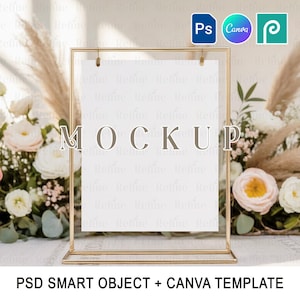Puede incluir: Un soporte de letrero de metal dorado con una inserción de papel blanco, que muestra la palabra "MOCKUP". El letrero está colocado sobre un fondo de flores y hierba de la pampa. El texto "PSD SMART OBJECT + CANVA TEMPLATE" está en la parte inferior.