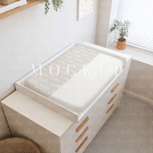 May include: A white changing table with three drawers and wooden handles. The changing pad has a light beige and white pattern. The image includes a small plant in a terracotta pot and a decorative wall art.