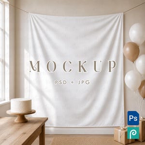 May include: A white fabric backdrop hanging on a string, featuring the centered text MOCKUP in a serif font, with the smaller text PSD + JPG below it. A white cake on a wooden stand sits on a table, with beige and white balloons to the right.