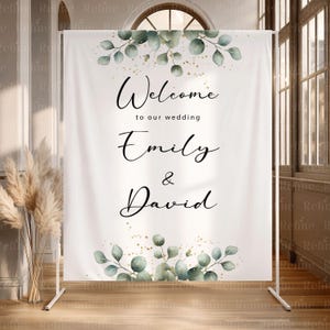 May include: A white wedding welcome sign with the names Emily & David. The sign has elegant script lettering and is decorated with watercolor eucalyptus leaves and gold flecks. The sign is displayed on a white metal stand.