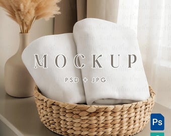 May include: A woven basket filled with two folded white towels. The basket sits on a neutral surface, with a vase of dried pampas grass and a window in the background. The word "MOCKUP" is displayed on the towels.