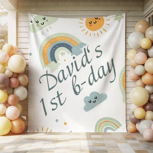 K&ouml;nnte beinhalten: Ein wei&szlig;es Stoffbanner mit Regenbogen-, Sonnen- und Wolkenmotiven und dem Text "David's 1st b-day" in t&uuml;rkisfarbener Schrift. Das Banner ist mit pastellfarbenen Luftballons auf beiden Seiten dekoriert.