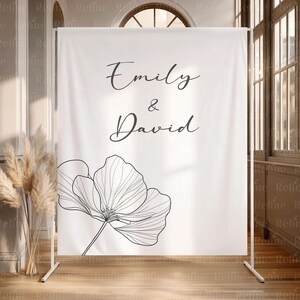 May include: A white backdrop with the names "Emily & David" in elegant script, accompanied by a large, black and white floral illustration. The backdrop is supported by a white metal frame, suitable for a wedding or event.
