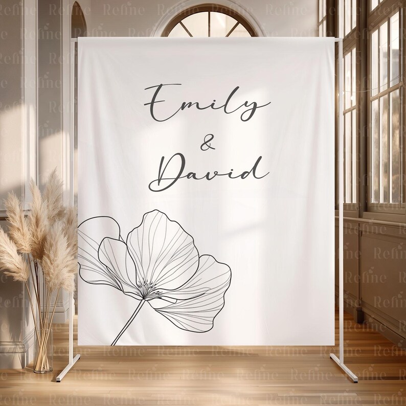 May include: A white backdrop with the names "Emily & David" in elegant script, accompanied by a large, black and white floral illustration. The backdrop is supported by a white metal frame, suitable for a wedding or event.