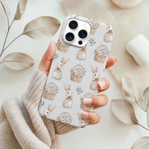 May include: A white phone case featuring a repeating pattern of Easter bunnies, baskets of flowers, and blue floral accents. The case has a glossy finish and a protective design, perfect for a smartphone.
