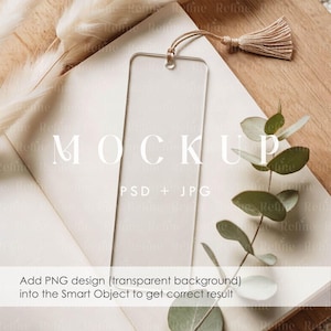 Puede incluir: Un marcapáginas de acrílico transparente con una borla beige, sobre una superficie blanca con la palabra "MOCKUP" en blanco. También es visible el texto "PSD + JPG". La imagen incluye ramitas de hojas verdes y plantas secas.