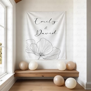 K&ouml;nnte beinhalten: Ein wei&szlig;es Banner mit den Namen "Emily & David" in eleganter Schrift und einem schwarz-wei&szlig;en Blumendesign. Mehrere beige Luftballons liegen auf dem Boden und eine Holzbank. Das Banner h&auml;ngt an einer wei&szlig;en Wand.