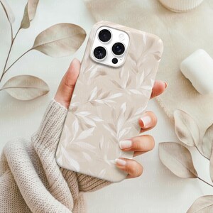 May include: A beige phone case with a repeating pattern of white and light brown leaves. The case is held in a hand, and the phone's camera lenses are visible. The overall aesthetic is soft and natural.