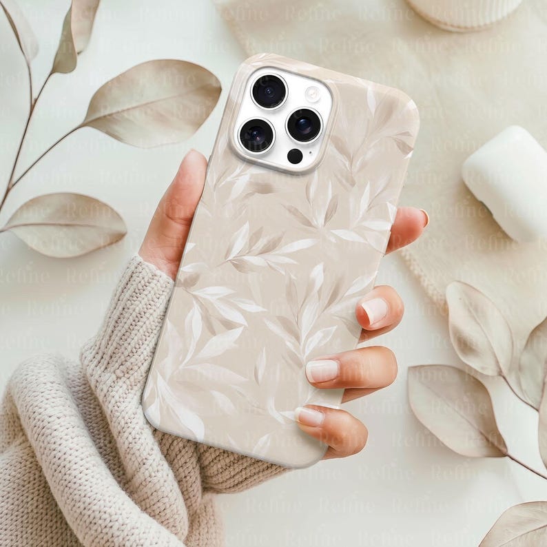 May include: A beige phone case with a repeating pattern of white and light brown leaves. The case is held in a hand, and the phone's camera lenses are visible. The overall aesthetic is soft and natural.