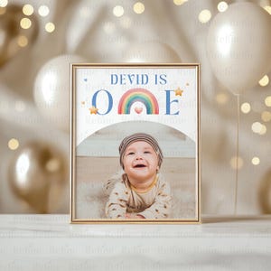 May include: A blue and white printable sign with a rainbow and stars. The sign says "DEVID IS ONE" and has a space for a photo. The photo shows a baby boy smiling.