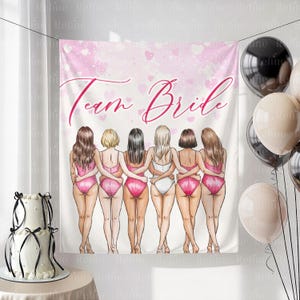 May include: A white banner with the text "Team Bride" in pink script, featuring an illustration of six women in pink swimsuits. Balloons and a two-tiered cake with black ribbons are also visible.
