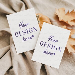 May include: Two white rectangular cards with the text "Your DESIGN here" in purple script, resting on a beige fabric. A small pumpkin and dried leaves are in the background, creating a fall theme.