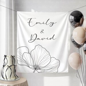 May include: A white backdrop with the names "Emily & David" in elegant script, accompanied by a large floral outline. A two-tiered cake with black ribbons and balloons in neutral tones complete the scene.