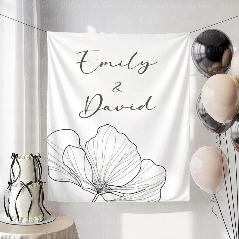 May include: A white backdrop with the names "Emily & David" in elegant script, accompanied by a large floral outline. A two-tiered cake with black ribbons and balloons in neutral tones complete the scene.