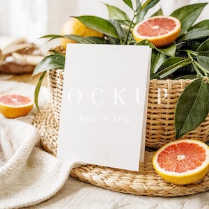 May include: A blank white card mockup stands in front of a woven basket filled with green leaves and sliced grapefruit. The basket sits on a woven placemat, with a soft, beige fabric in the foreground. The scene evokes a fresh, natural aesthetic.