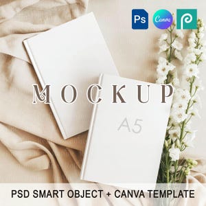 May include: Two white A5 notebooks with the word "MOCKUP" overlaid on the image. The notebooks are on a beige fabric with white flowers. The text "PSD SMART OBJECT + CANVA TEMPLATE" is at the bottom.