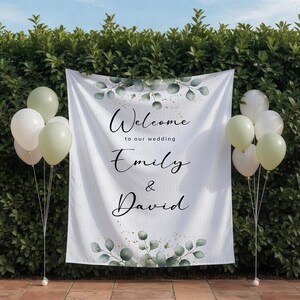 May include: A white wedding banner with the words "Welcome to our wedding Emily & David" in elegant script. The banner is decorated with green eucalyptus leaves and gold accents. Balloons in white and green hues flank the banner.