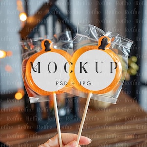 Puede incluir: Dos piruletas naranjas con forma de calabaza sobre palitos de madera, cada una en un envoltorio de plástico transparente. Las piruletas tienen un círculo blanco en el centro con la palabra "MOCKUP" y "PSD + JPG". El fondo está borroso.