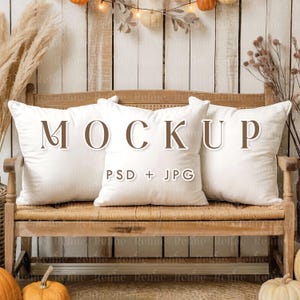 Puede incluir: Tres cojines cuadrados blancos con la palabra "MOCKUP" en letras marrones están colocados en un banco de madera. El texto "PSD + JPG" está debajo de la palabra "MOCKUP". El fondo incluye calabazas, follaje otoñal y luces de cadena.
