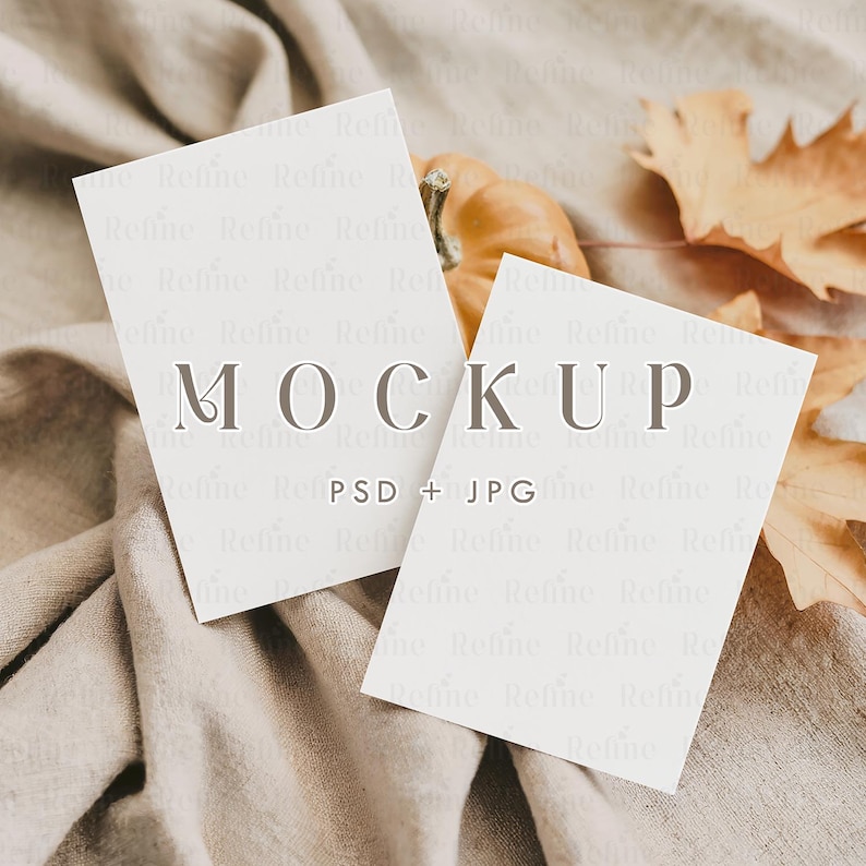 May include: Two white rectangular cards with the word "MOCKUP" printed on them, along with "PSD + JPG". The cards are resting on a beige fabric, with a small pumpkin and dried leaves in the background. The overall theme is autumn.